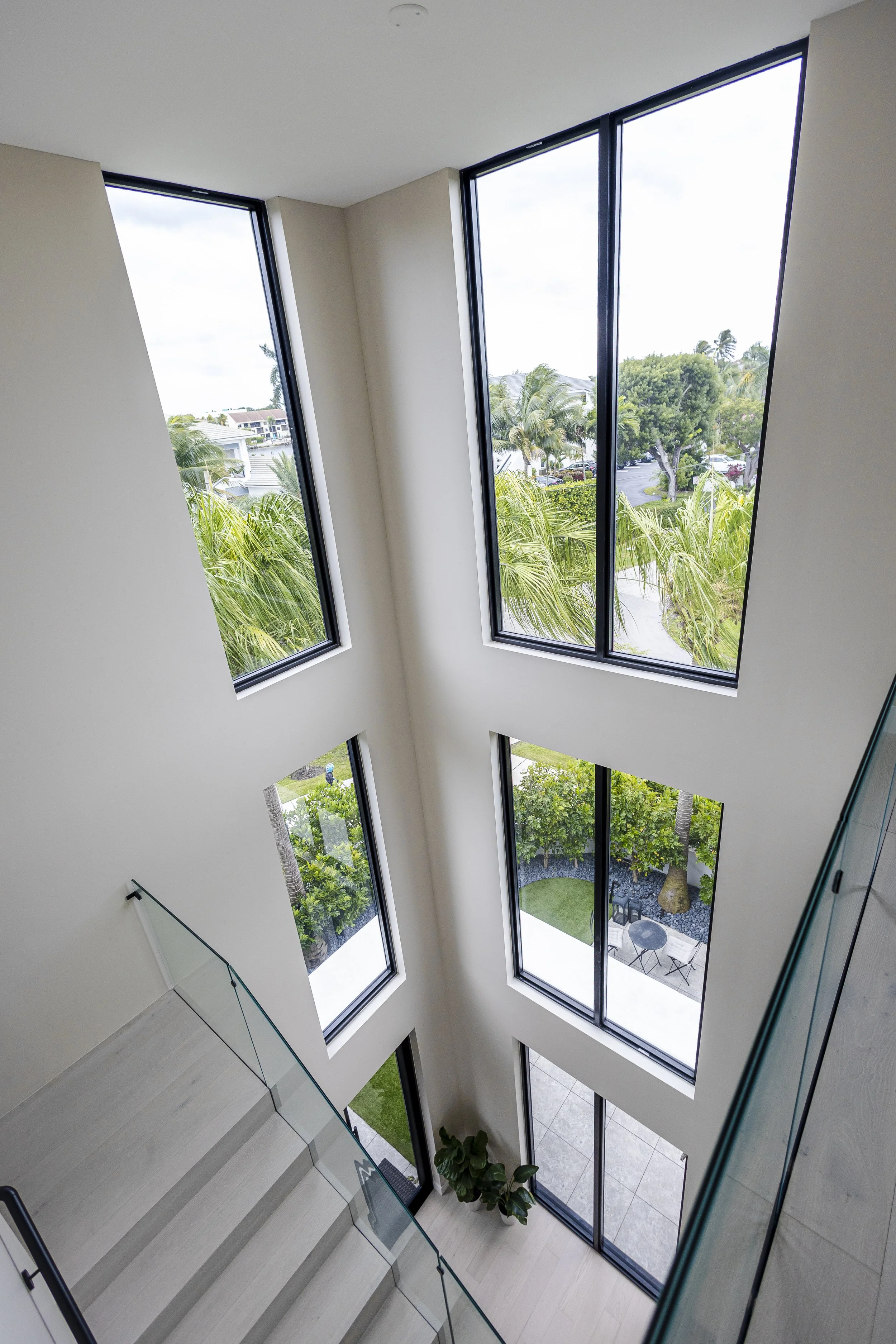 A view from the top of a stairway looking down through a glass railing at a modern interior with large, tall windows showing lush green palm trees and outdoor seating area.