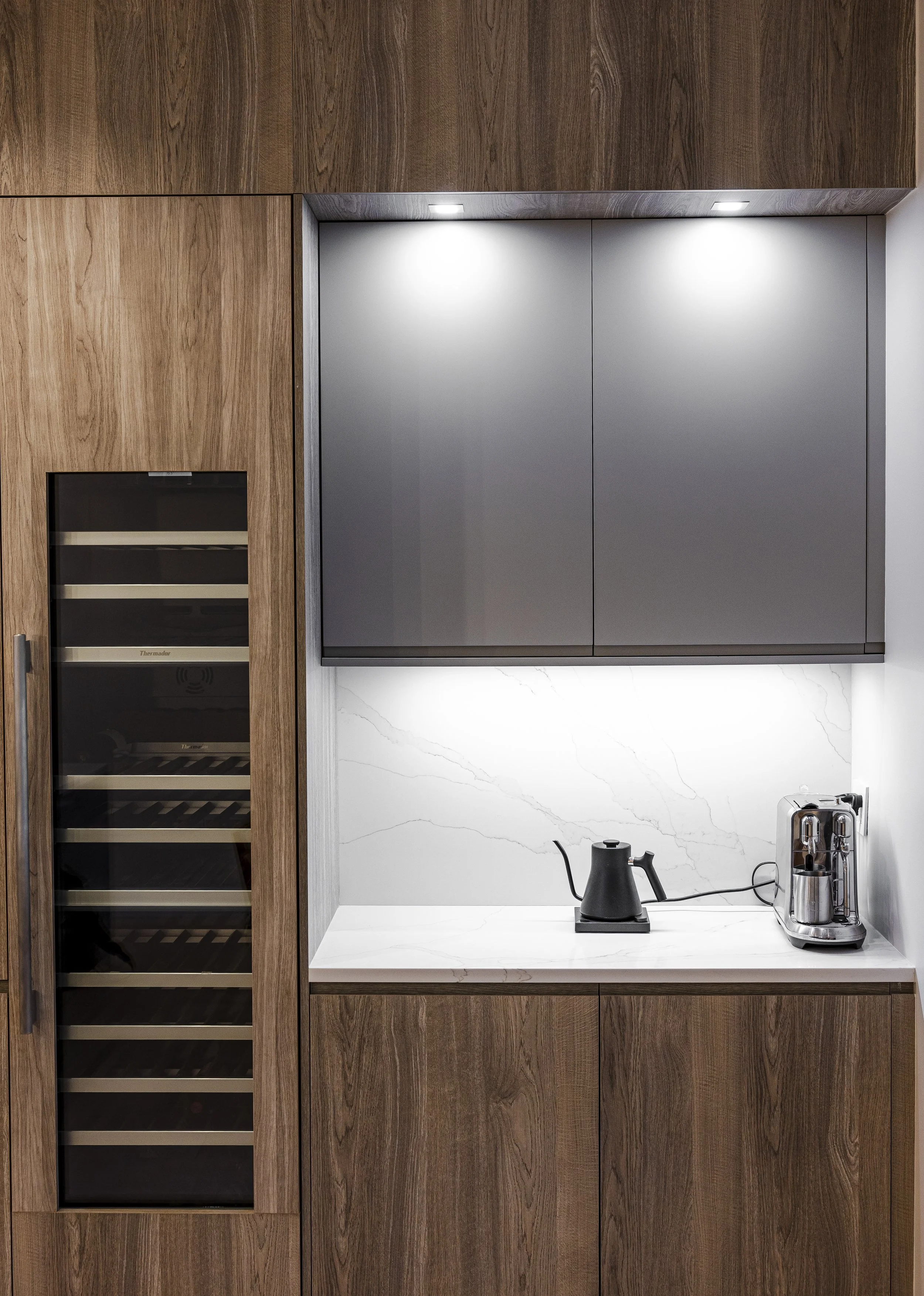A modern kitchen nook with wooden cabinets, a white marble countertop with gray veining, and a marble backsplash. There is a black teapot on an electric stove, and a silver coffee machine on the right side.