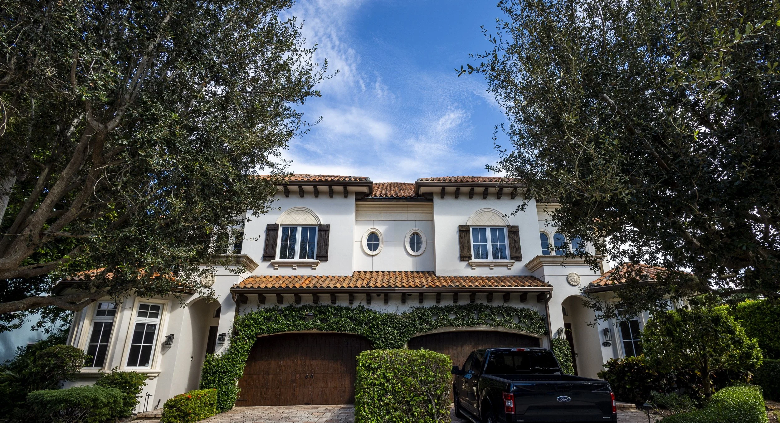 Large white house with terracotta roof, surrounded by trees and green bushes, and a black Ford truck parked in front.