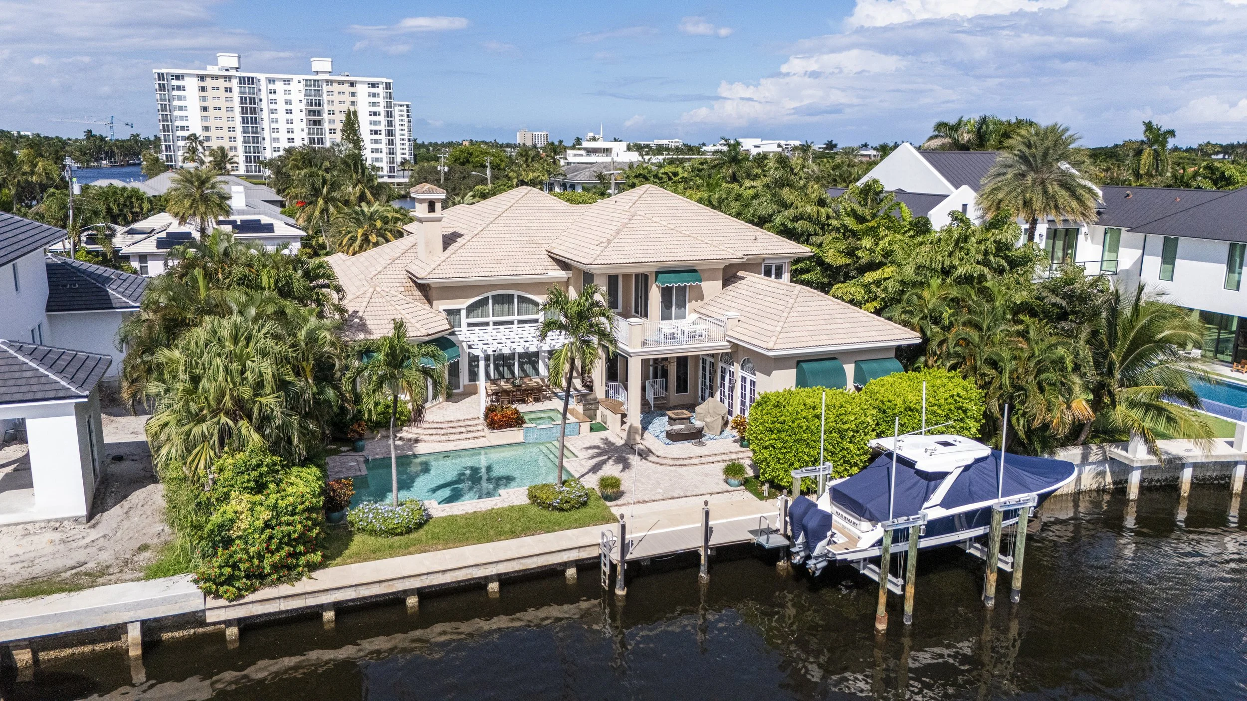 A large beach house with a tiled roof, pool, and balcony, located by a waterway with a boat docked at the pier. Surrounded by greenery and neighboring houses, with high-rise buildings in the background.