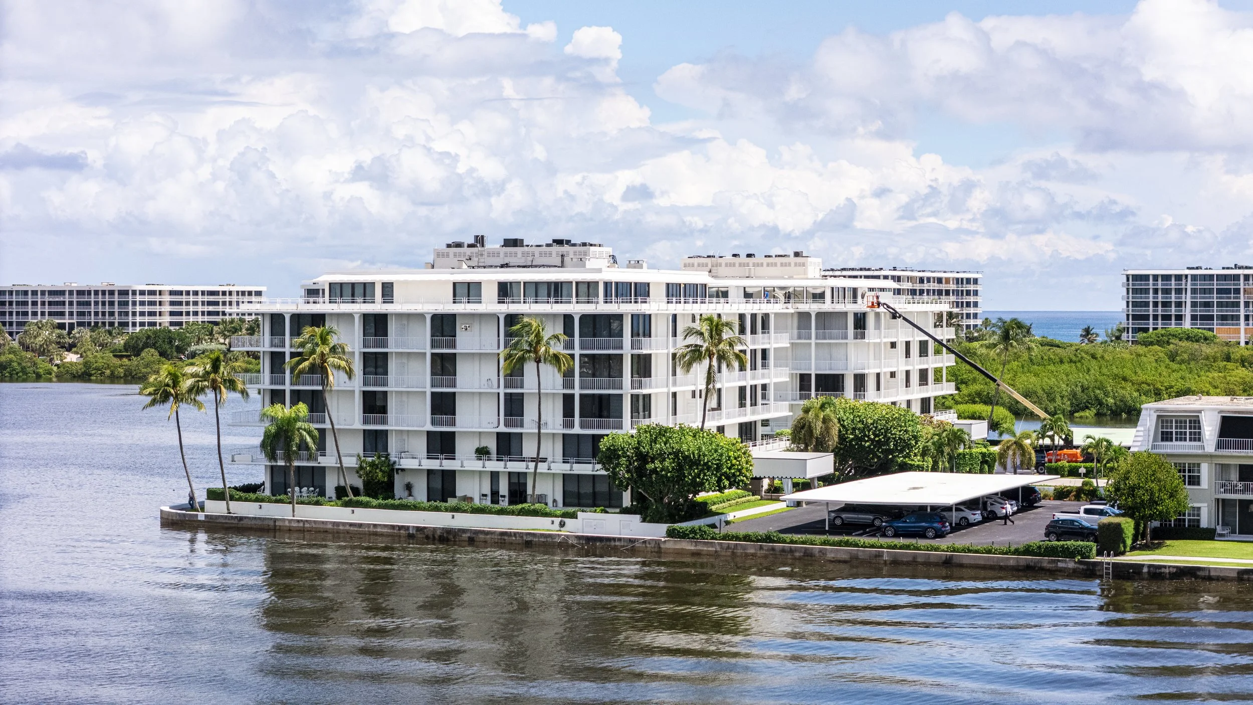 High-rise residential building by water with palm trees, parking area, and a construction crane.