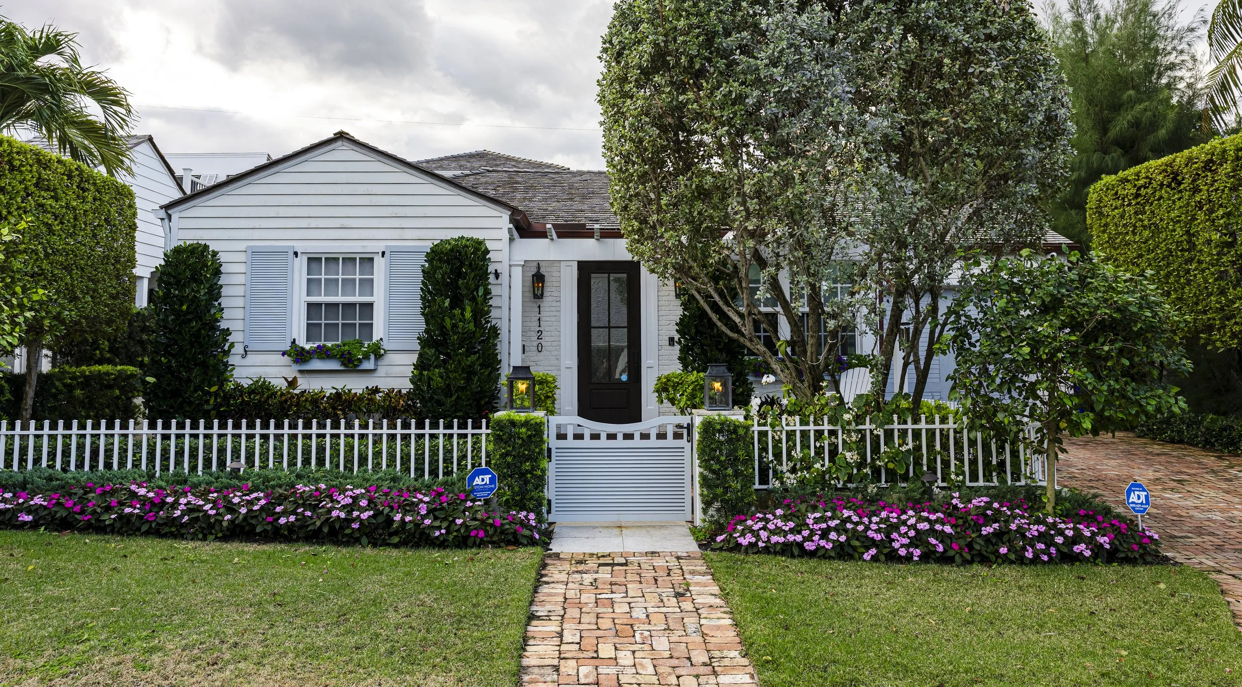 A charming white house with a black front door, surrounded by a white picket fence, lush green shrubs, trees, and colorful flowers in a landscaped yard.
