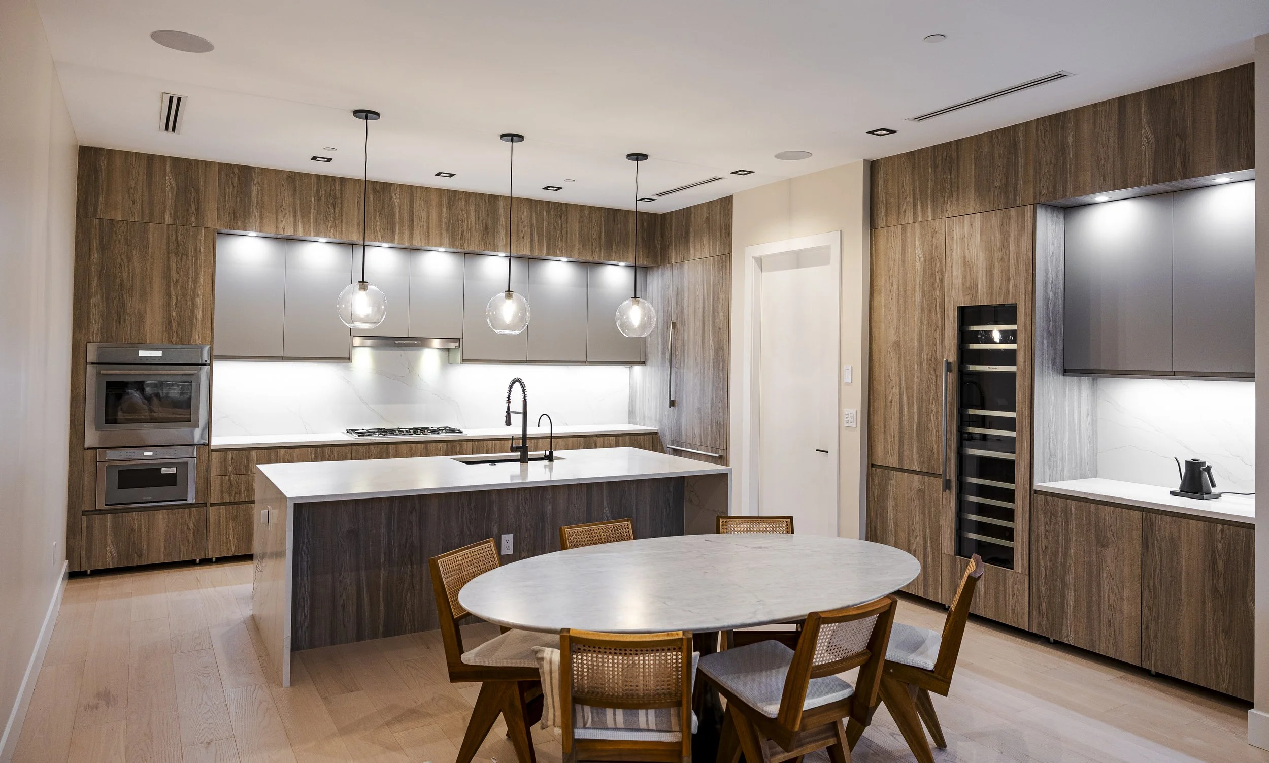 Modern kitchen with wooden cabinets, white marble backsplash, island with black sink, pendant lights, and a round dining table with six chairs.