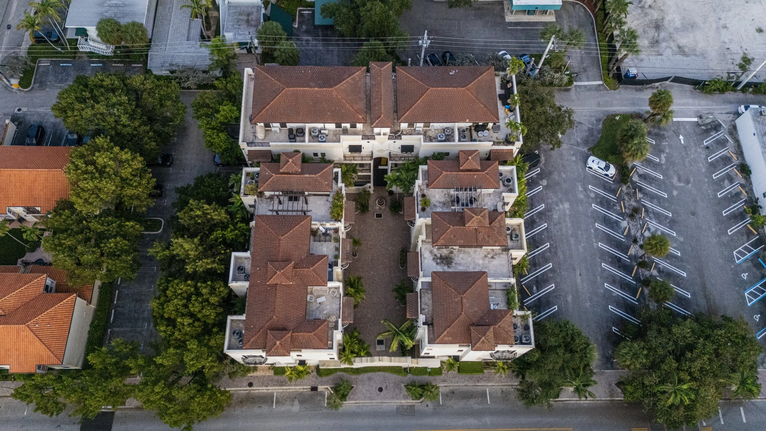 Aerial view of a residential apartment complex with red-tiled roofs, surrounded by trees, parking lots, and adjacent streets.