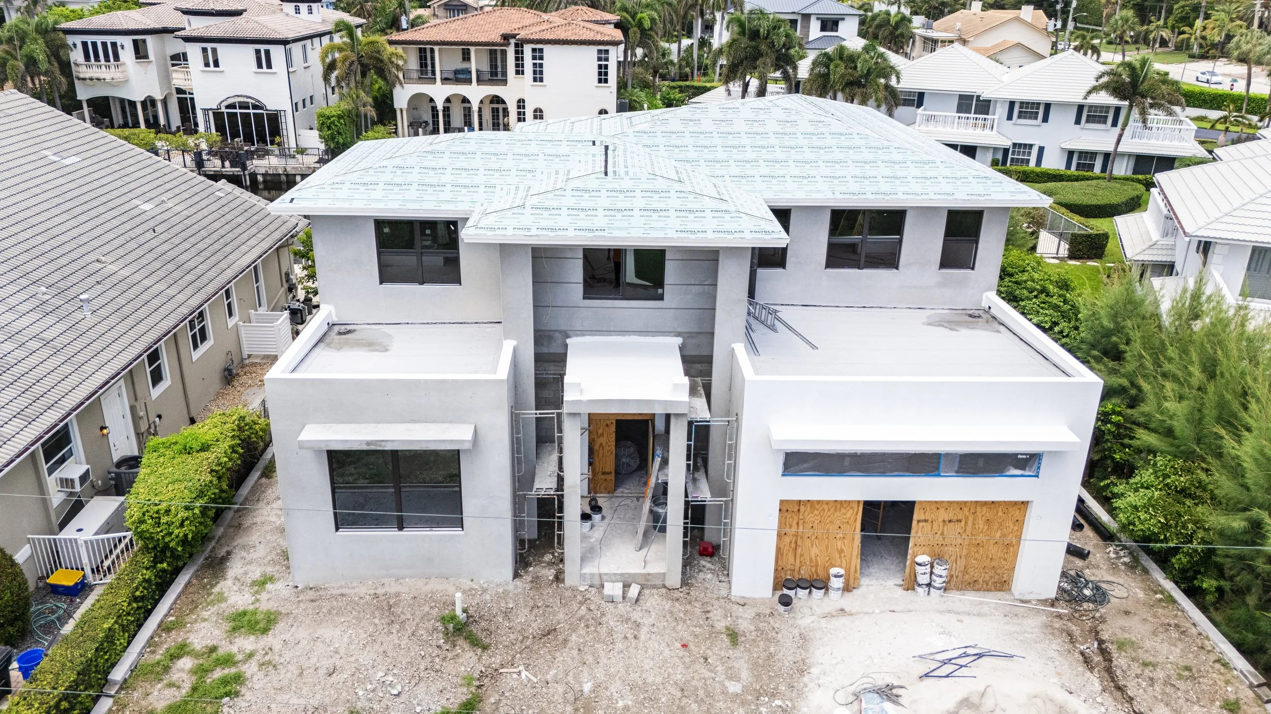 An aerial view of a two-story house under construction with a gray stucco exterior, flat roof, and large windows, surrounded by neighboring houses in a residential neighborhood.