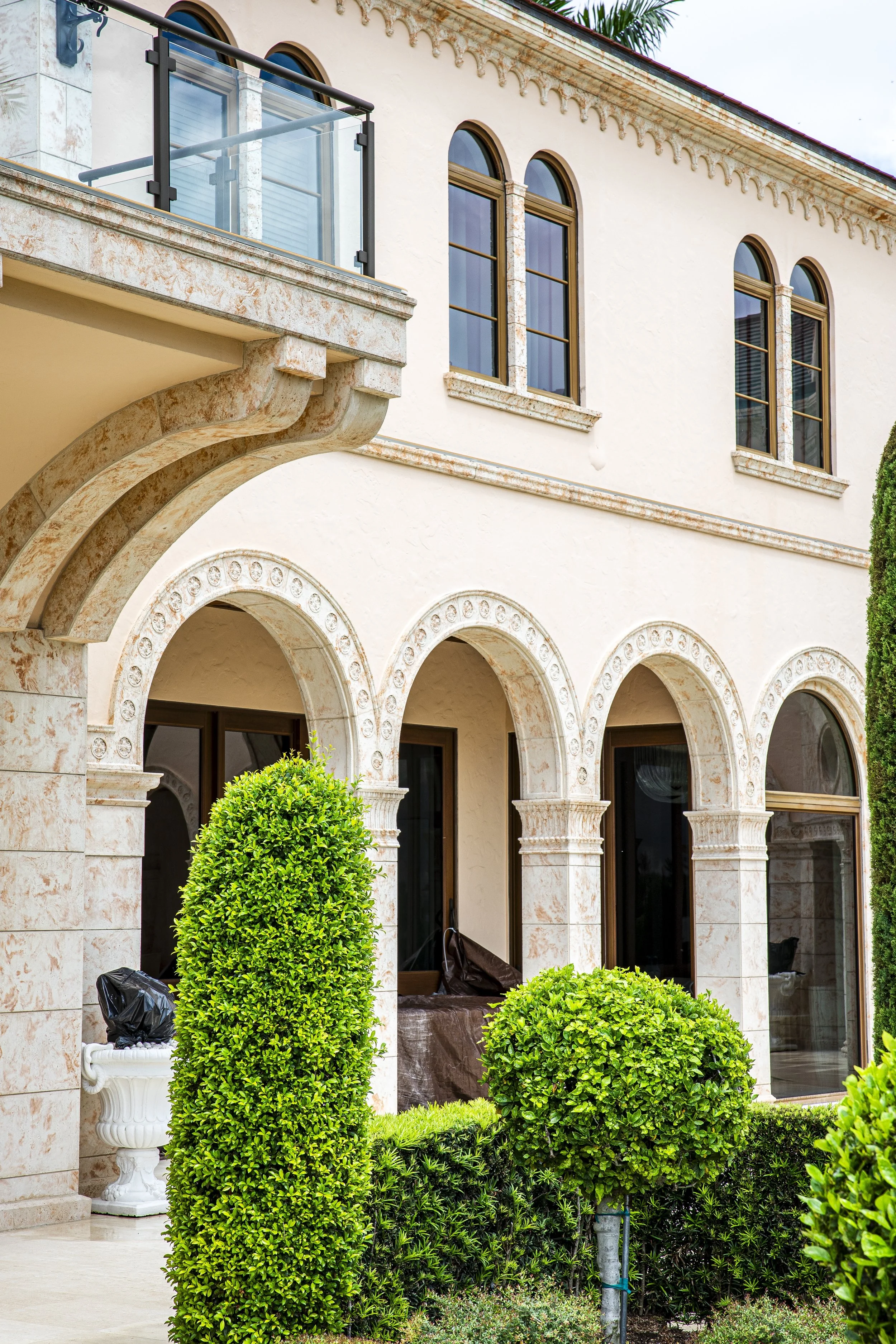 A beige stucco house with arched windows and decorative stonework, with manicured green bushes in the front yard.