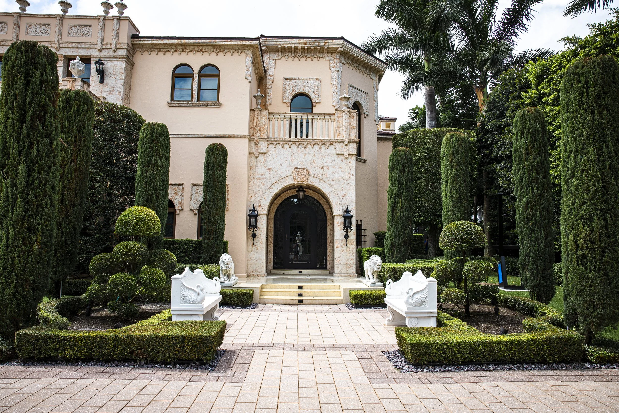 Front view of a luxurious mansion with manicured landscaping, including tall cypress trees, spherical bushes, and statues of lions and swans, with steps leading to a black front door.