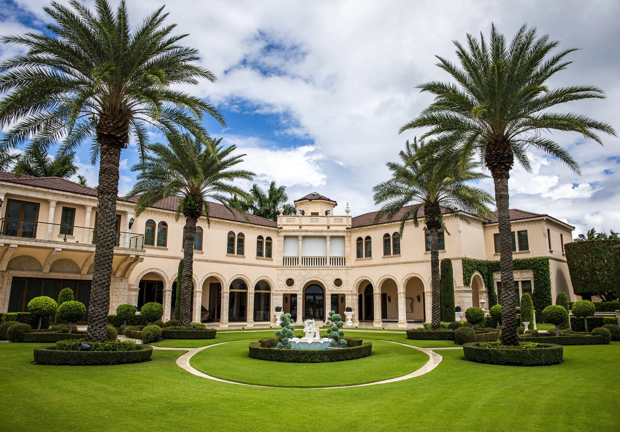 Luxurious mansion with a manicured lawn, palm trees, a fountain, and a cloudy sky.