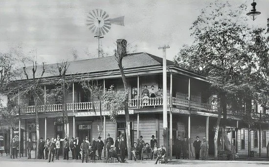 Black and white photo of a two-story building with a balcony and people standing on the sidewalk and on the balcony, possibly from the early 20th century.