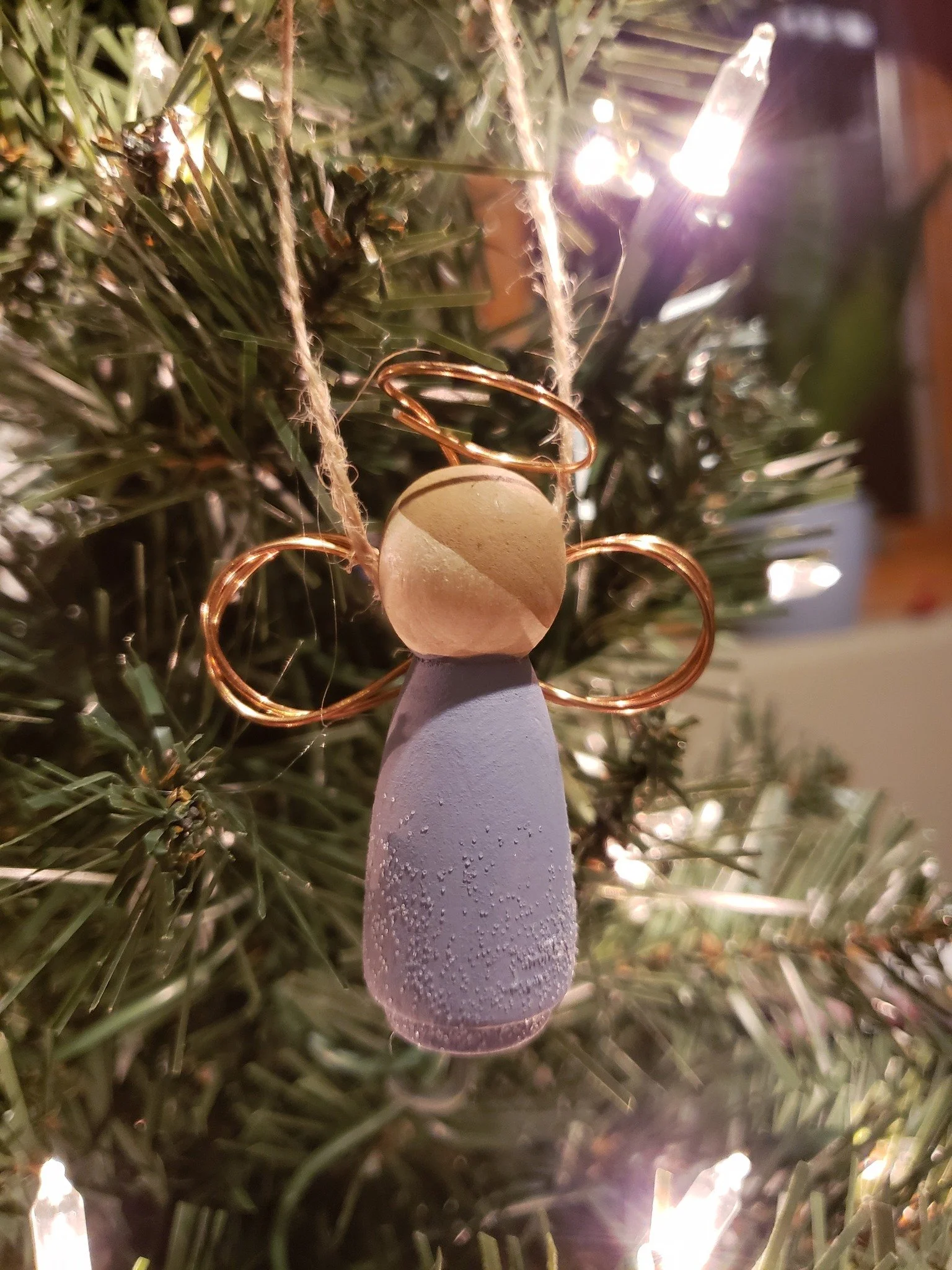 A Christmas tree ornament that resembles a snowman with a blue body and a beige head, equipped with copper wire loops for arms and a loop for hanging. The ornament is decorated with condensation, indicating it's cold, and the tree is adorned with whi