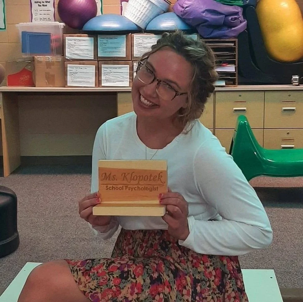 Woman smiling, holding a wooden nameplate that reads, 'Ms. Klopotek, School Psychologist,' sitting on a classroom mat with gym equipment and supplies in the background.