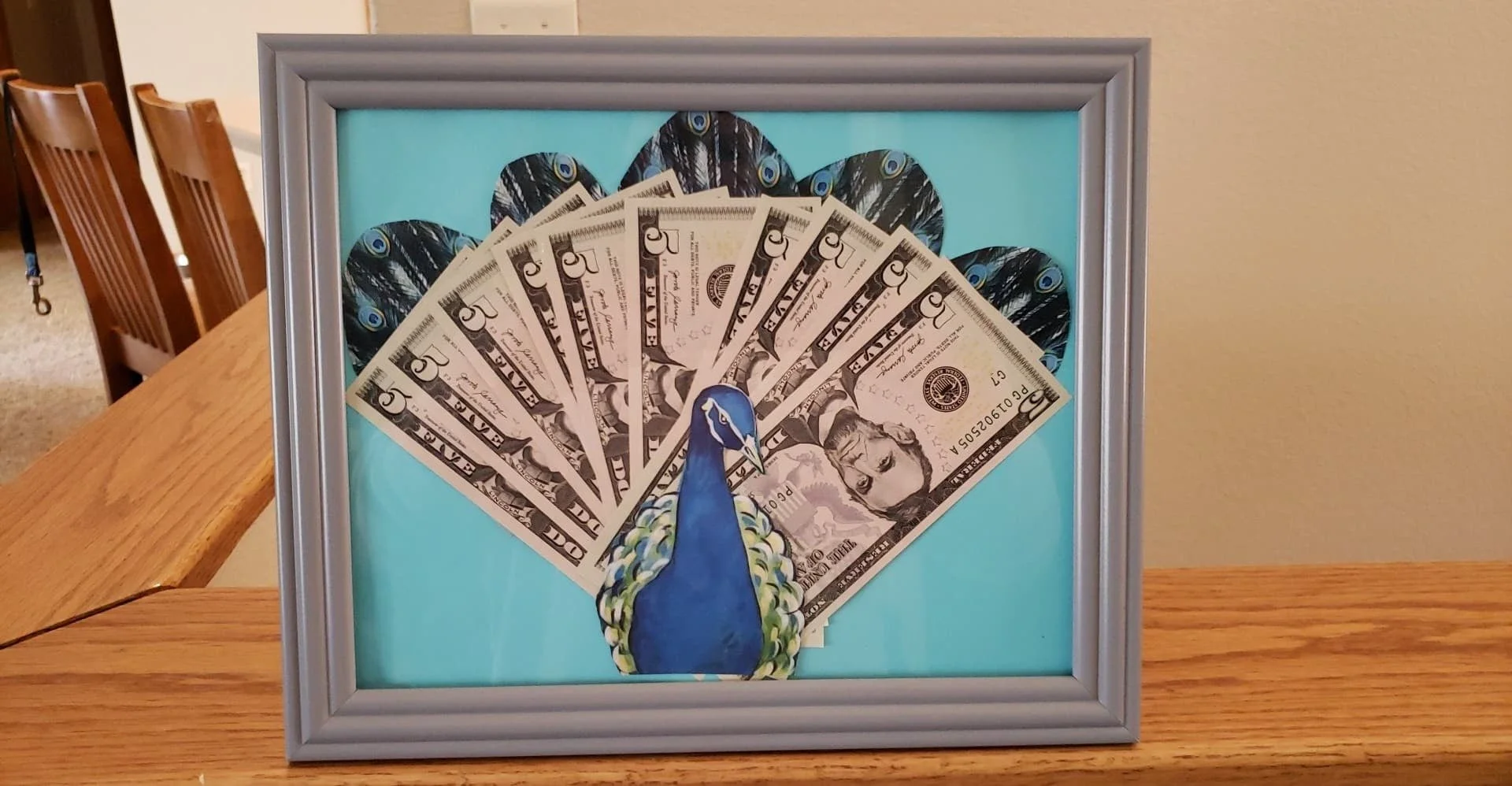 A framed artwork of a peacock with dollar bills as its tail feathers, displayed on a wooden table.
