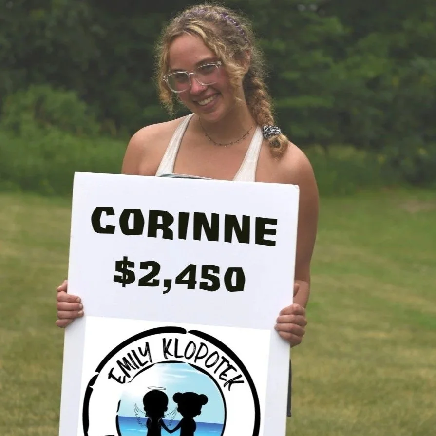 A woman with glasses and curly hair holding a sign reading 'Corinne $2,450' and a logo for 'Emily Klopotek' with a graphic of a child and an angel holding hands in front of a body of water.