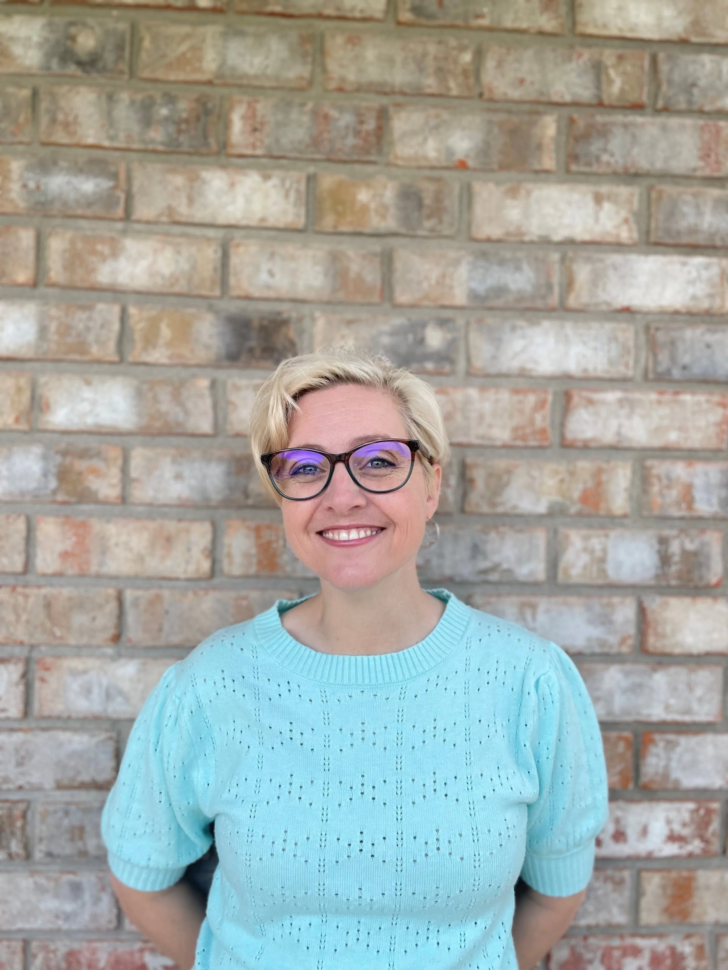 Smiling woman with blonde hair and glasses standing in front of a brick wall.