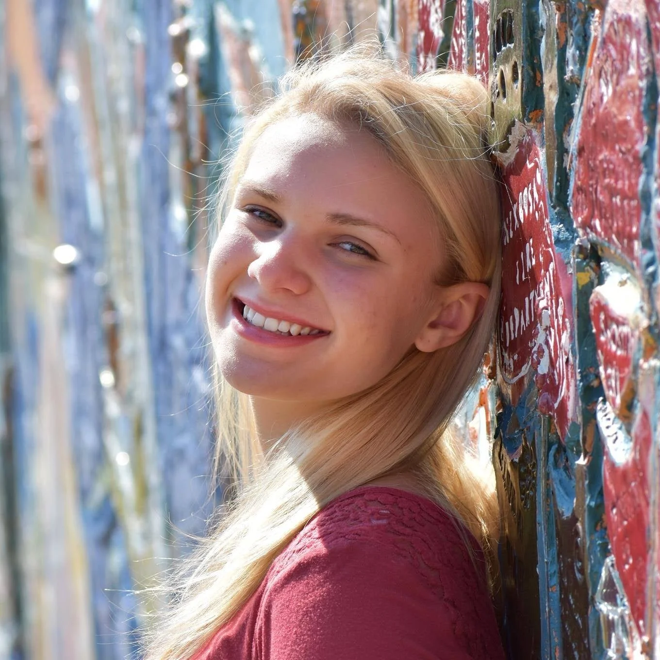 A young woman with long blonde hair smiling and leaning against a colorful, textured wall outdoors.