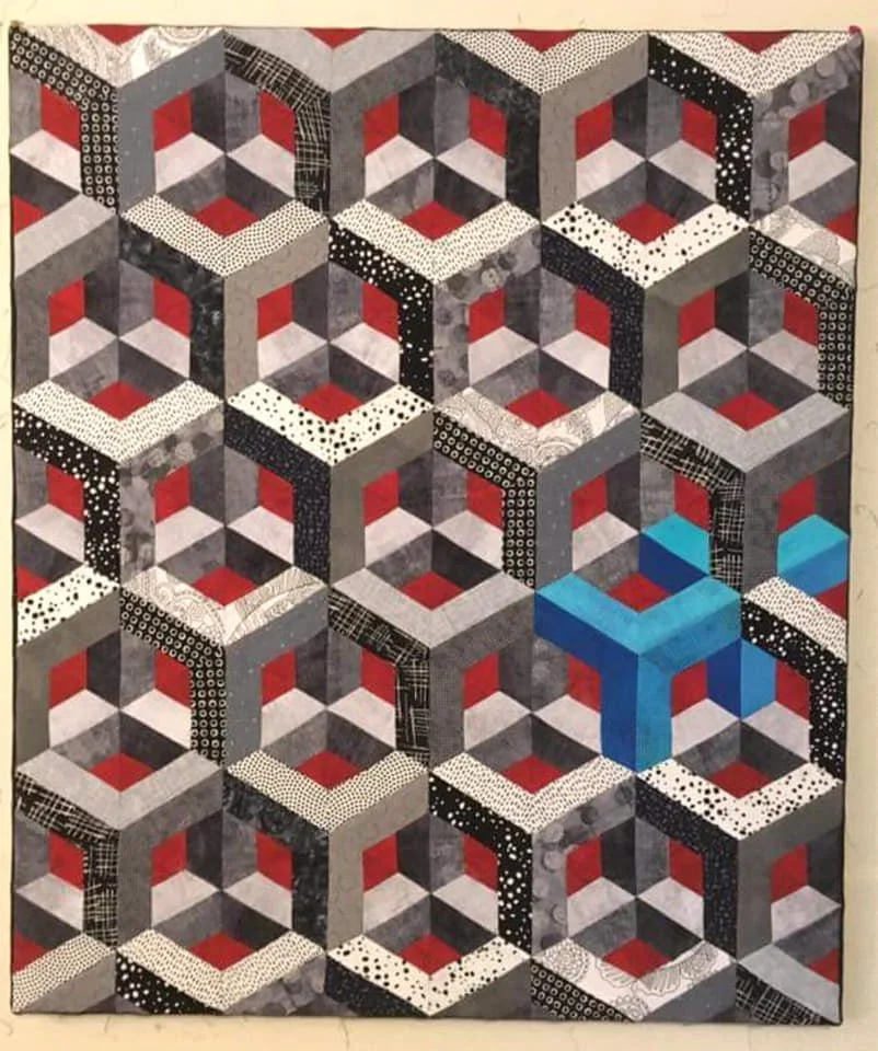 A quilt with a repeating pattern of three-dimensional cubes created by interlocking hexagons. The quilt features colors of red, gray, black, white, and a blue cube in the center.
