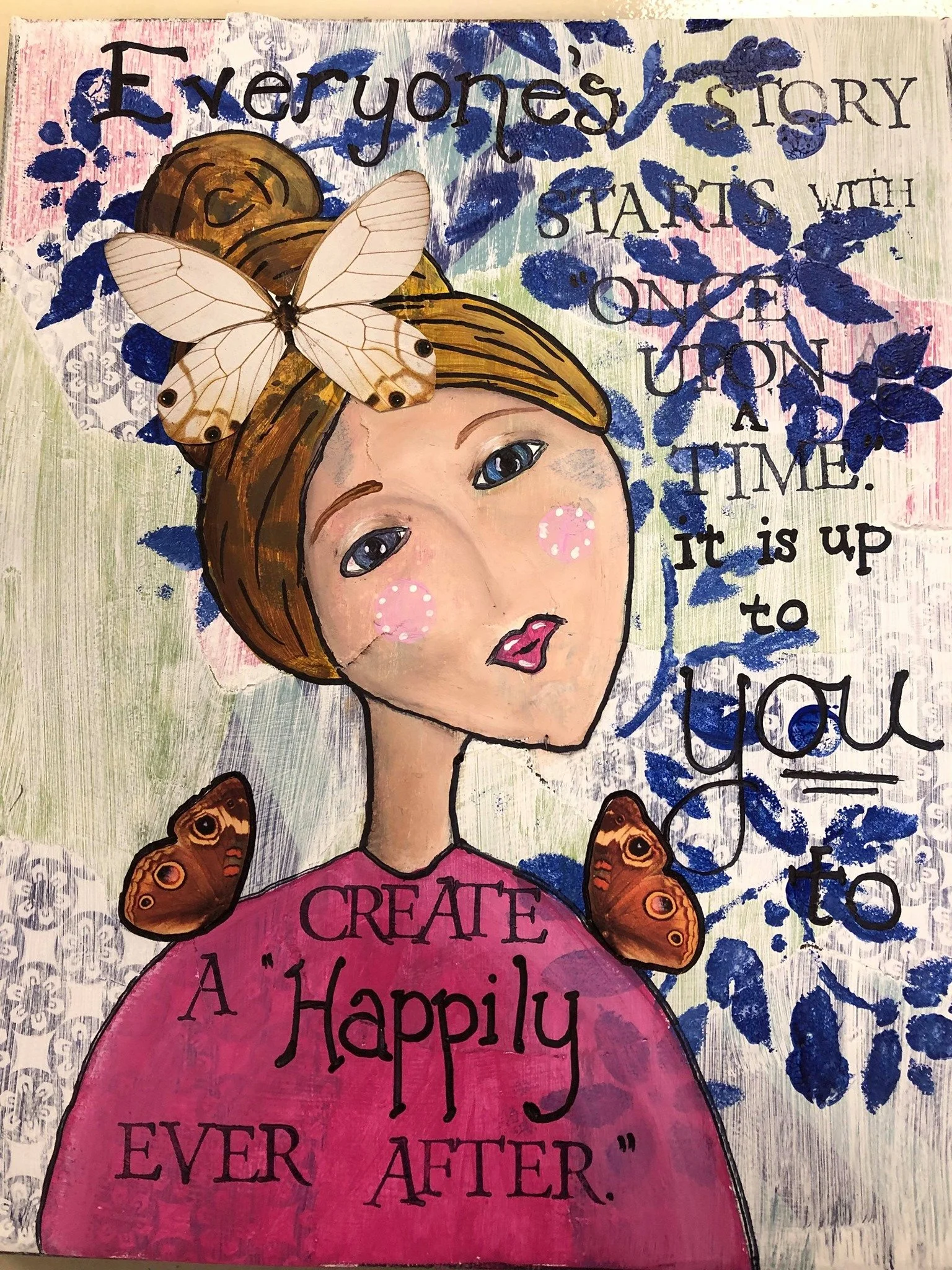 Colorful art piece featuring a woman with brown hair, blue eyes, pink cheeks, wearing a pink shirt with the words "CREATE A Happily EVER AFTER." Two butterflies are on her shoulders, and a white butterfly is on her hair. Blue leaves and pink and whit