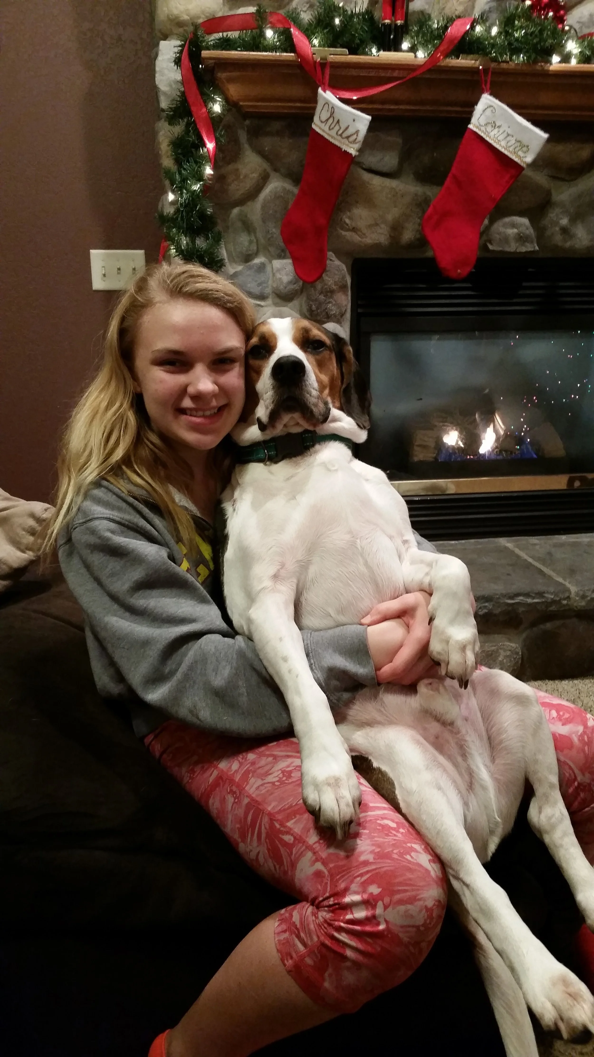 Girl with long blonde hair smiling while sitting on a couch, holding a large white and brown dog with floppy ears in front of a decorated fireplace with Christmas stockings and garland.