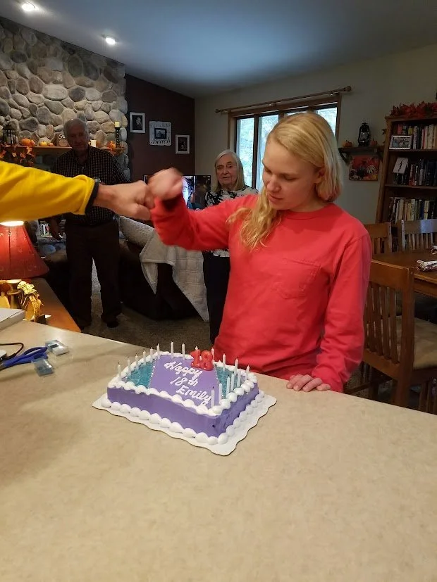 Young girl with blonde hair, wearing a red long-sleeve shirt, standing at a kitchen counter with a purple birthday cake that says "Happy 16th, Emily" in white icing, candles on the cake, and someone in a yellow shirt handing her a pink item. In the background, three older adults and a stone fireplace are visible inside a cozy home.
