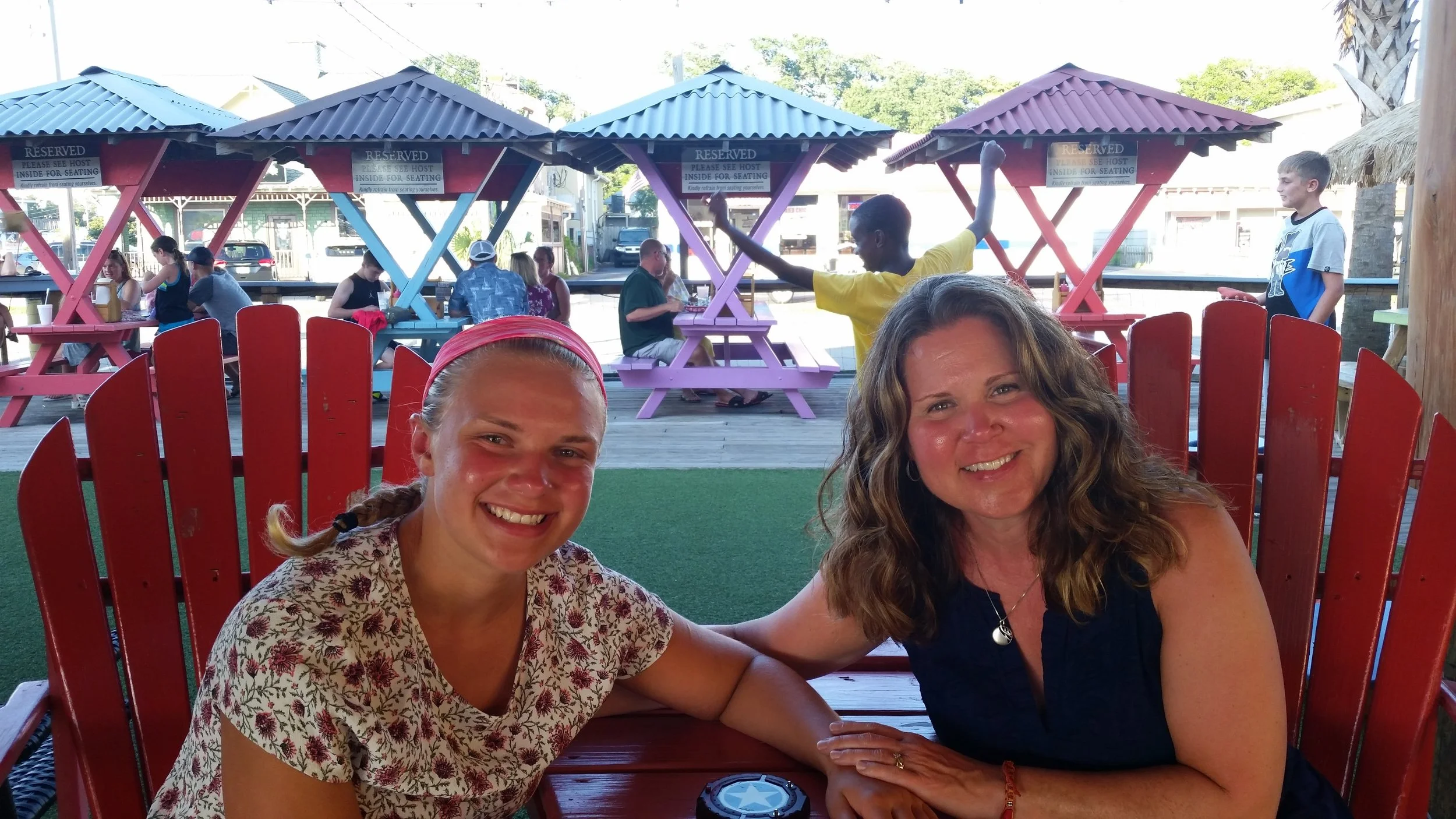 Two women sitting at a red table outdoors, smiling. In the background, there are colorful picnic shelters, people sitting and socializing, and a few children playing.
