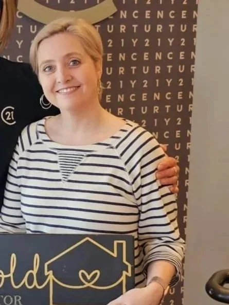 A woman with shoulder-length blonde hair, wearing a white and navy striped shirt, smiling and standing indoors near a dark background with text.