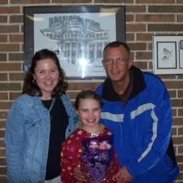 Three people, two women and a girl, smiling and standing together in front of a brick wall and framed pictures.