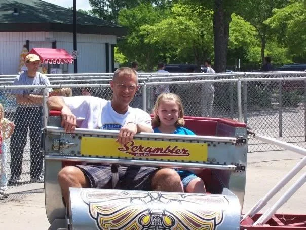 A man and a young girl sitting in a amusement park ride called 'Scrambler,' smiling at the camera, surrounded by a chain-link fence and other visitors.