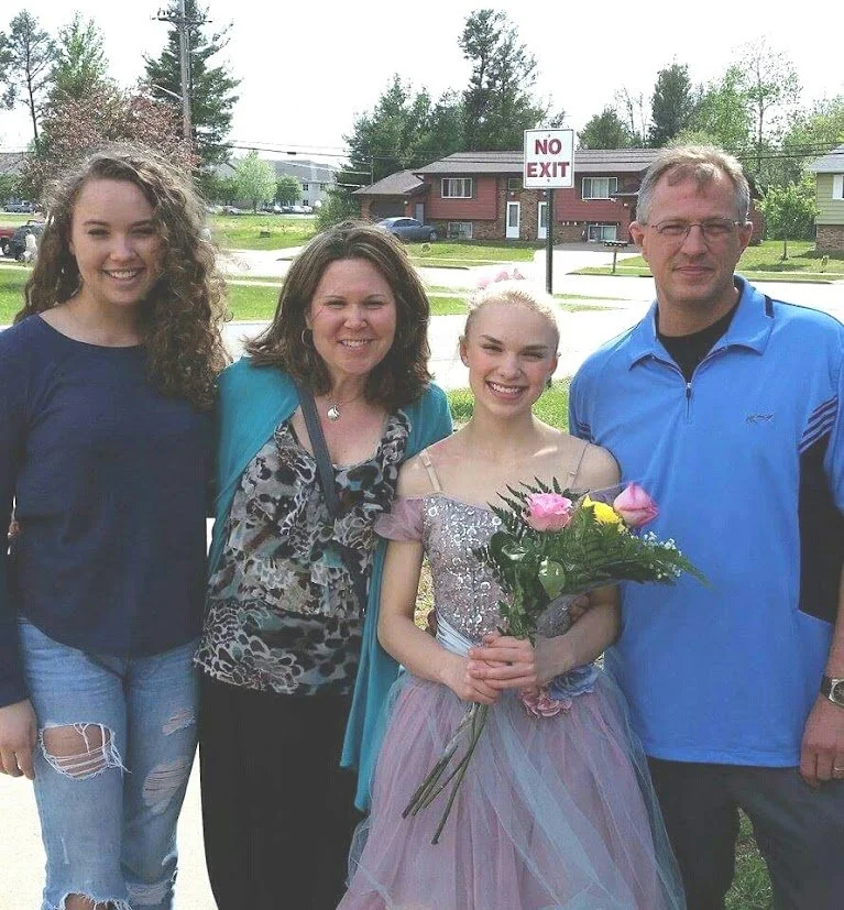 Group of four people outdoors, young girl in a pastel dress holding a bouquet of pink flowers, two women and one man smiling, residential neighborhood in background.