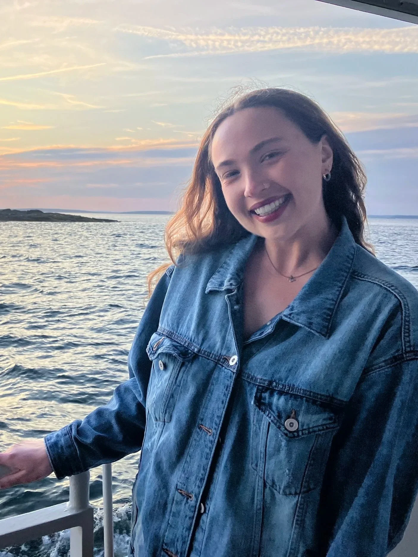 A woman with light skin and curly brown hair smiling on a boat at sunset, wearing a denim jacket with water and sky in the background.