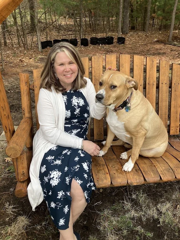 A woman sitting on a wooden bench outdoors with a large tan and white dog. The woman is smiling and wearing a navy blue dress with white floral pattern and a white cardigan. The dog is sitting next to her, looking forward, with a black collar.