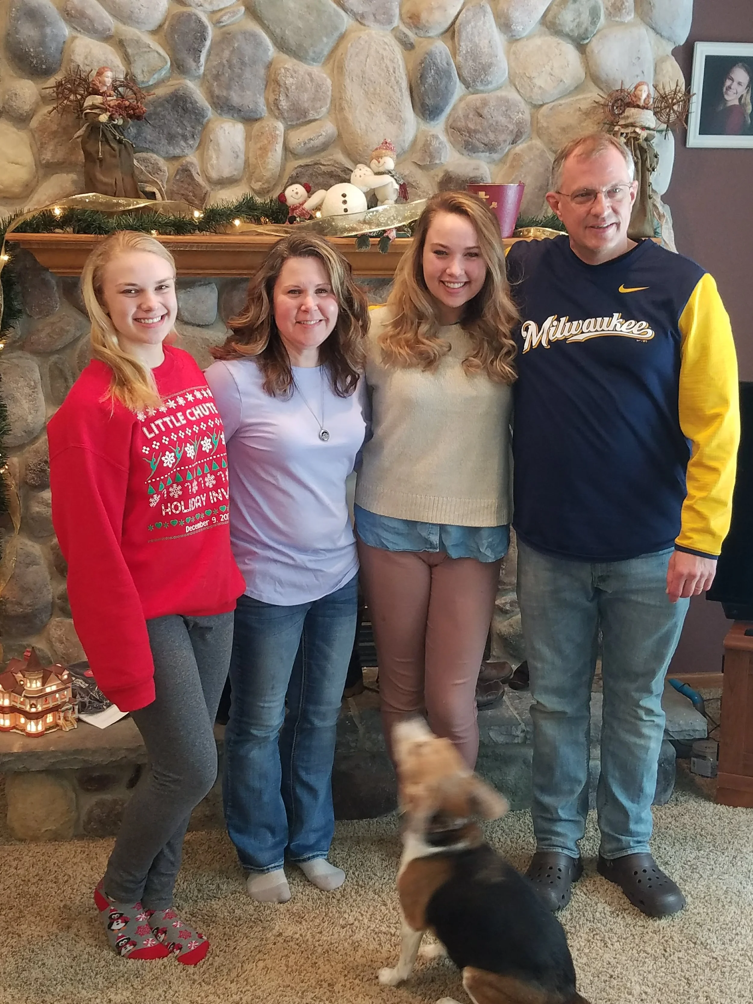 Four people, three women and one man, standing in front of a decorated stone fireplace, smiling for the photo. A small dog is in the foreground, blurred and looking upwards. The fireplace has holiday decorations including snowmen, snowflakes, and greenery.