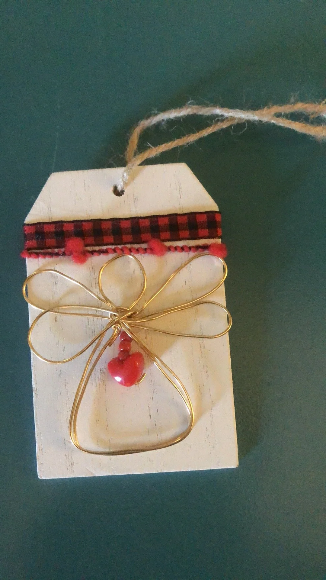 A decorative gift tag with a cream-colored wooden background, adorned with red and black checkered ribbon, red pom-pom trim, a gold wire bow, and red heart-shaped beads.