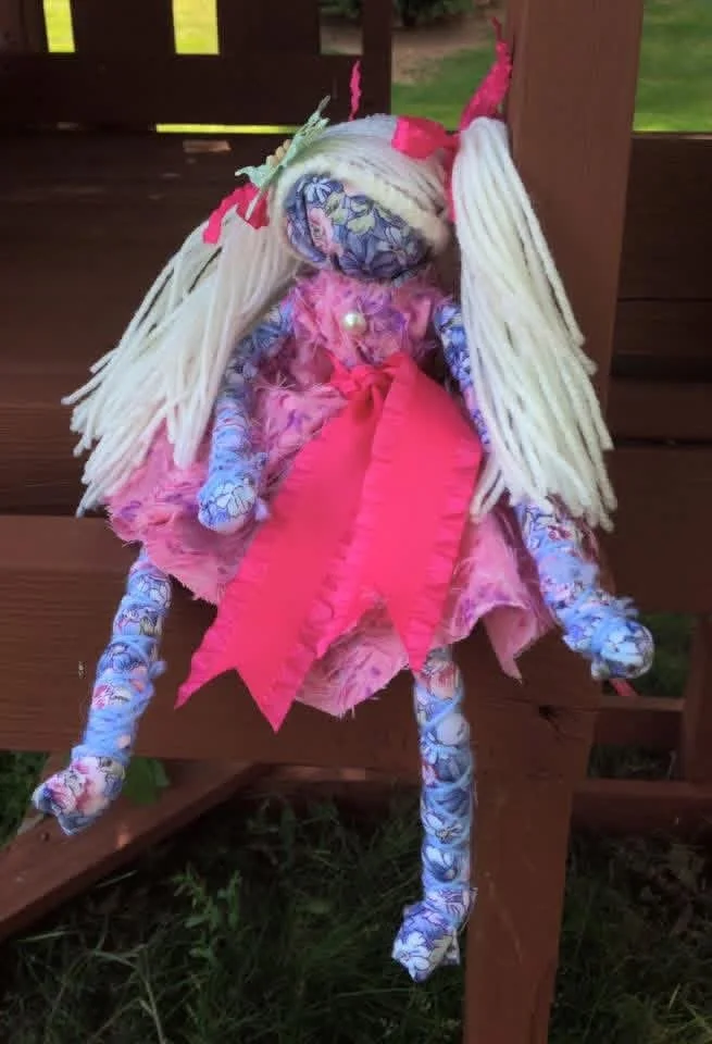 Colorful fabric doll dressed as a fairy with long white yarn hair, pink dress, and pink wings, sitting on a wooden bench outdoors.