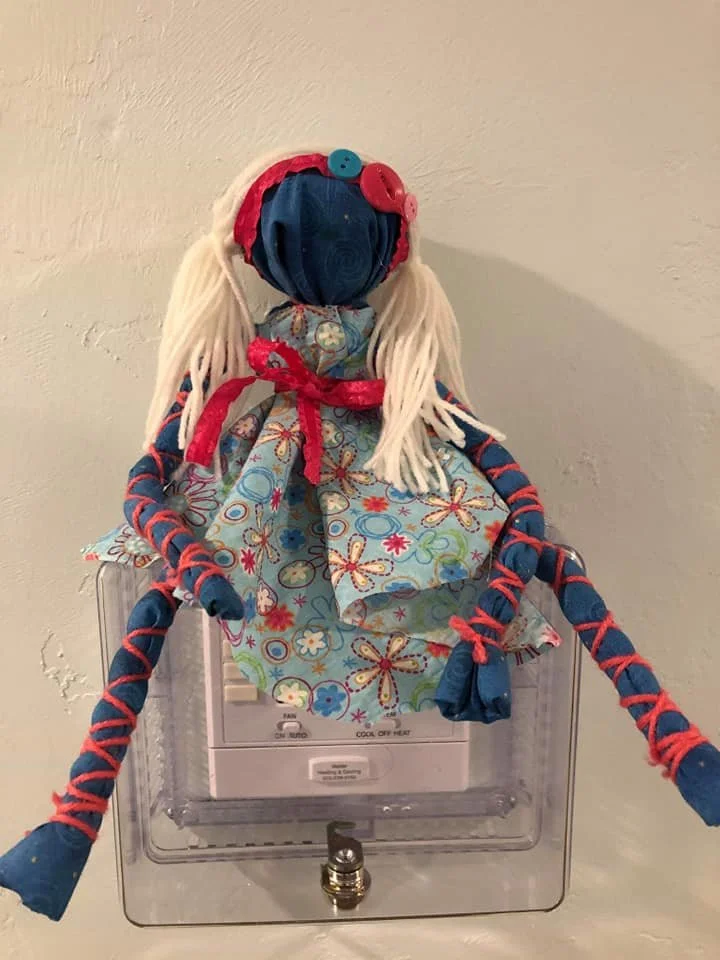 A handmade cloth doll with yarn hair, dressed in a floral dress and accessories, sitting on a fire alarm pull station.