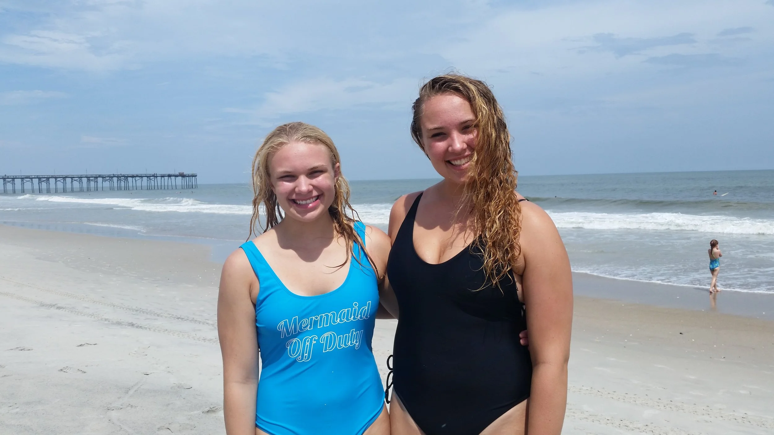 Two smiling women in swimsuits standing on a sandy beach near the ocean with a pier in the background.