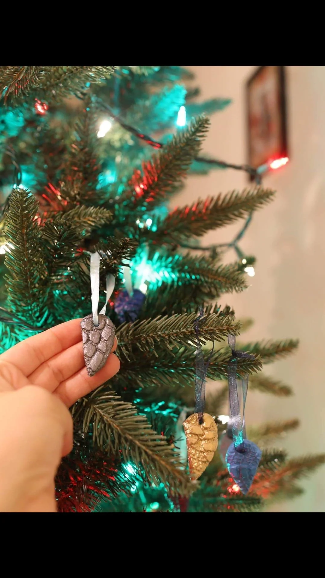 Close-up of a Christmas tree decorated with colorful lights and pinecone-shaped ornaments, with a hand holding a silver pinecone ornament in the foreground.