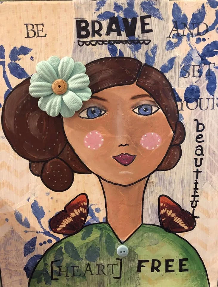 A colorful mixed media artwork of a girl's portrait with a floral accessory in her hair, surrounded by inspirational words like 'Be Brave,' 'And Beautiful,' and 'Heart Free' in a decorative, painted background.