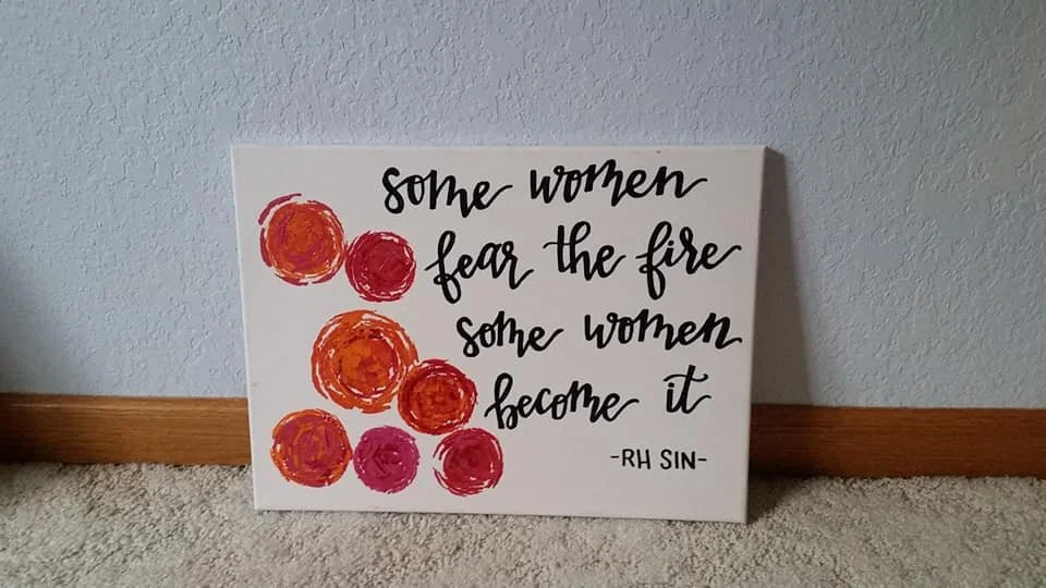 A decorative sign with a quote and painted red, orange, and pink circles. The quote reads, 'Some women fear the fire, some women become it,' attributed to RH Sin.
