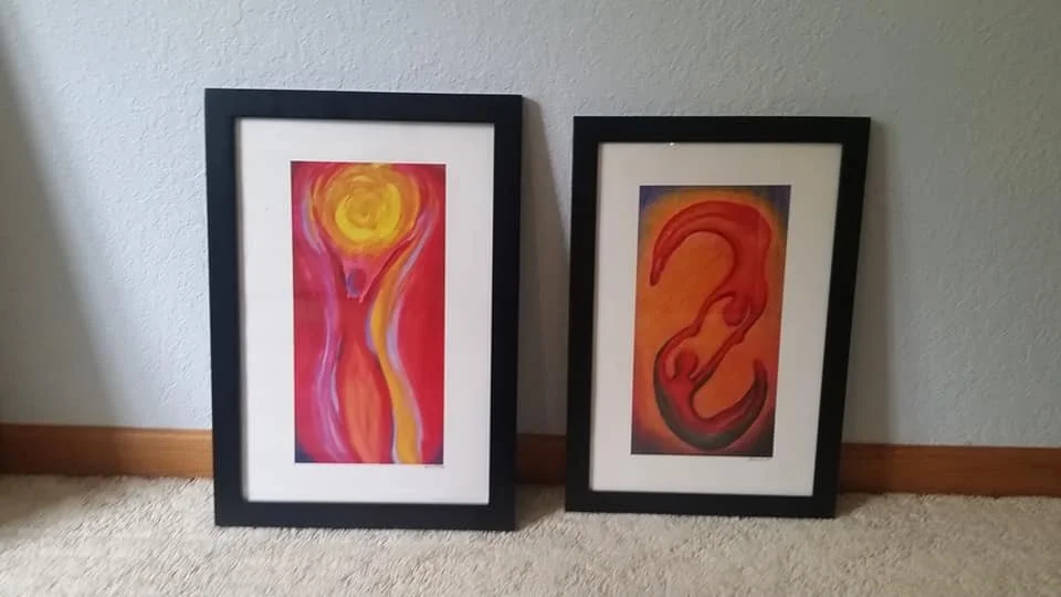 Two framed abstract paintings leaning against a wall on a beige carpeted floor. The left artwork features warm colors with a yellow sun and flowing shapes. The right artwork has a large, stylized red-orange swirl against a dark background.