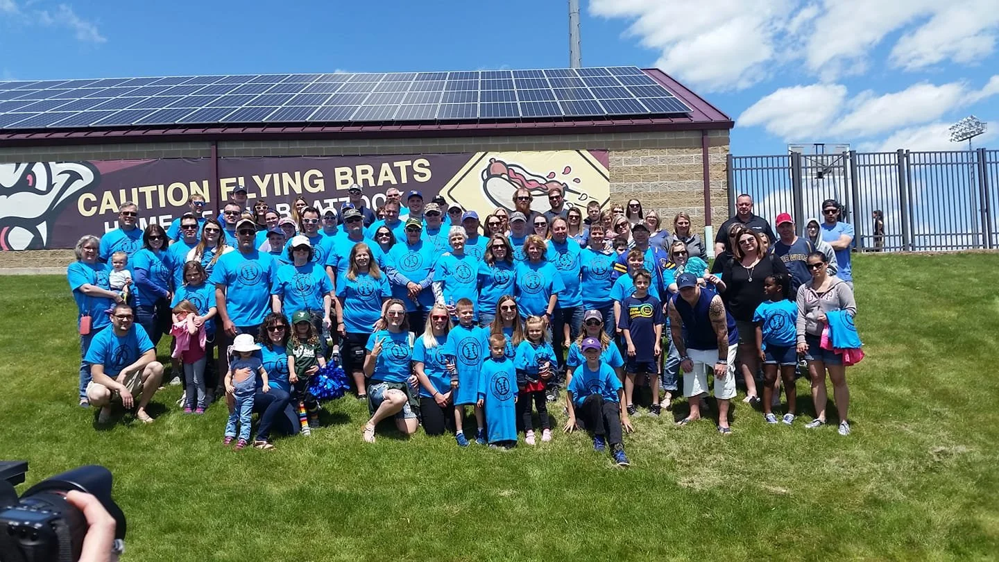 Group of people posing on a grassy field in front of a building with a sign that says "Caution Flying Brats." Many are wearing matching blue T-shirts with a logo, and some children are holding balloons or wearing hats. The sky is partly cloudy, and there is a camera in the lower left corner of the photo.