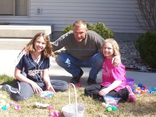 Father and two daughters Easter egg hunt outdoors in front of house with plastic eggs and basket.