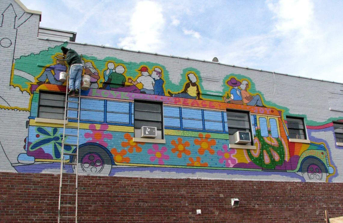 A mural of a colorful retro bus with flower motifs painted on a brick wall, with people sitting and relaxing depicted on the upper part of the bus.