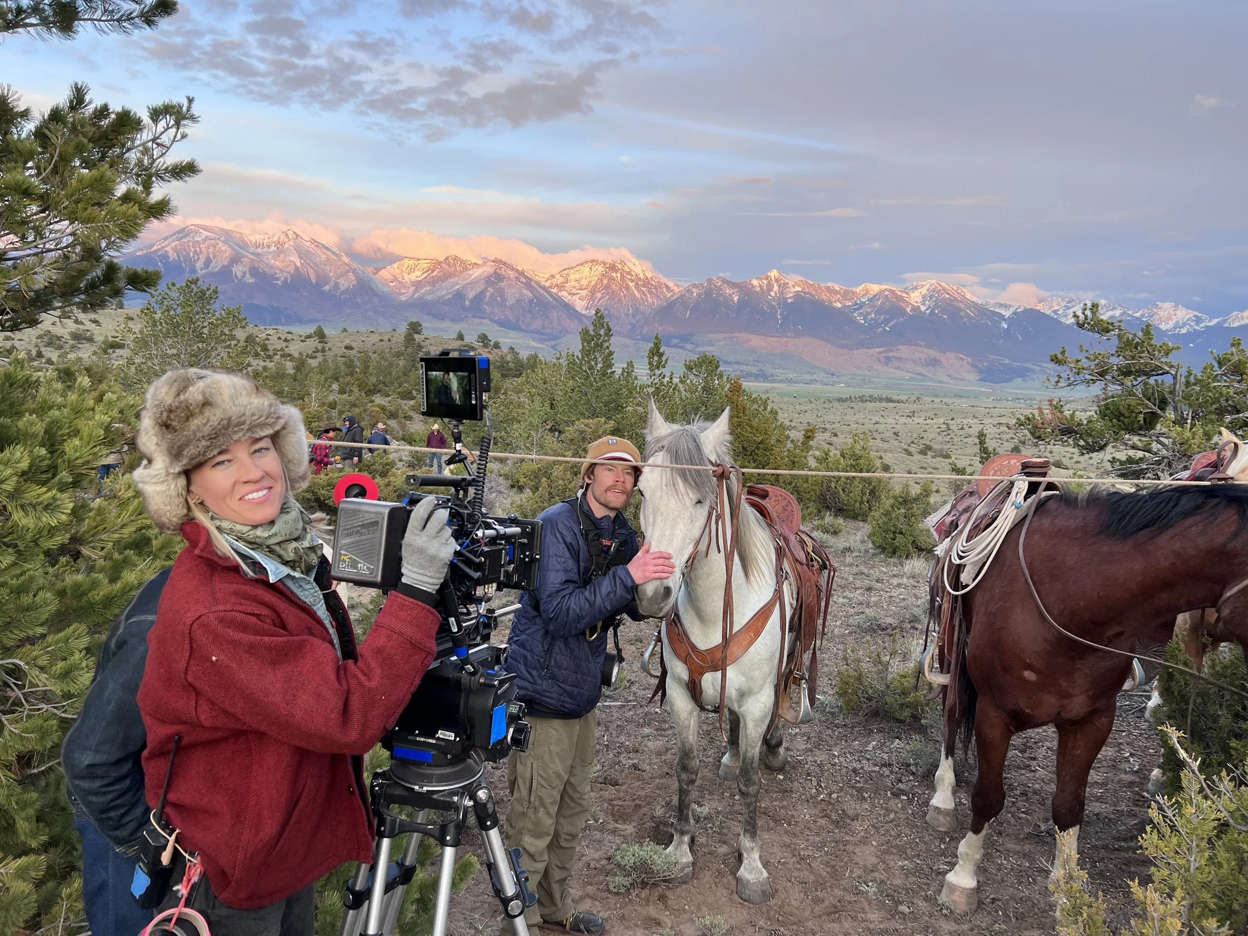 Two people filming or taking photos with a camera and tripod in a mountainous landscape with snow-capped peaks in the background, along with parked horses and greenery.
