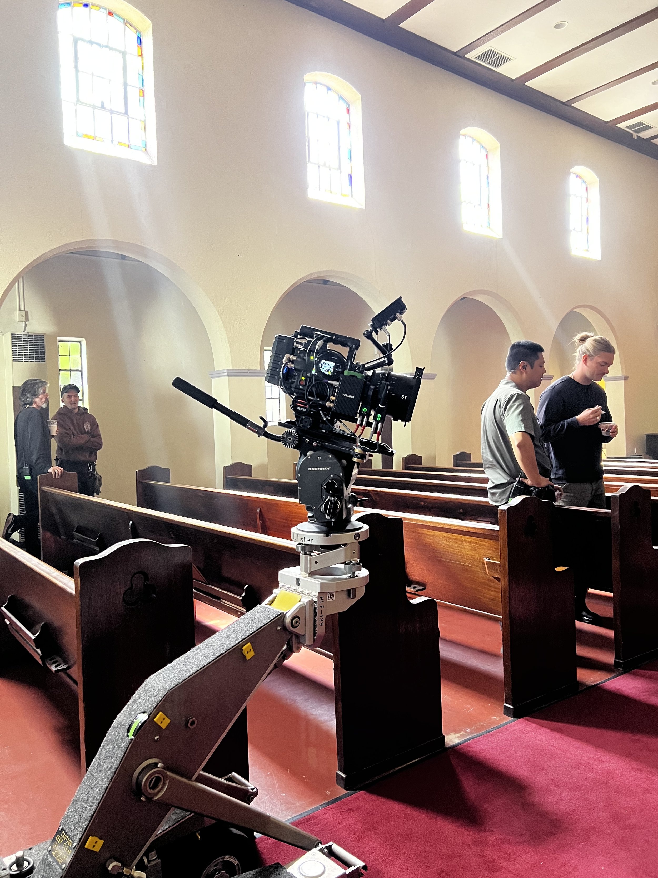 Film crew with camera on a dolly inside a church with wooden pews, stained glass windows, and light streaming in.