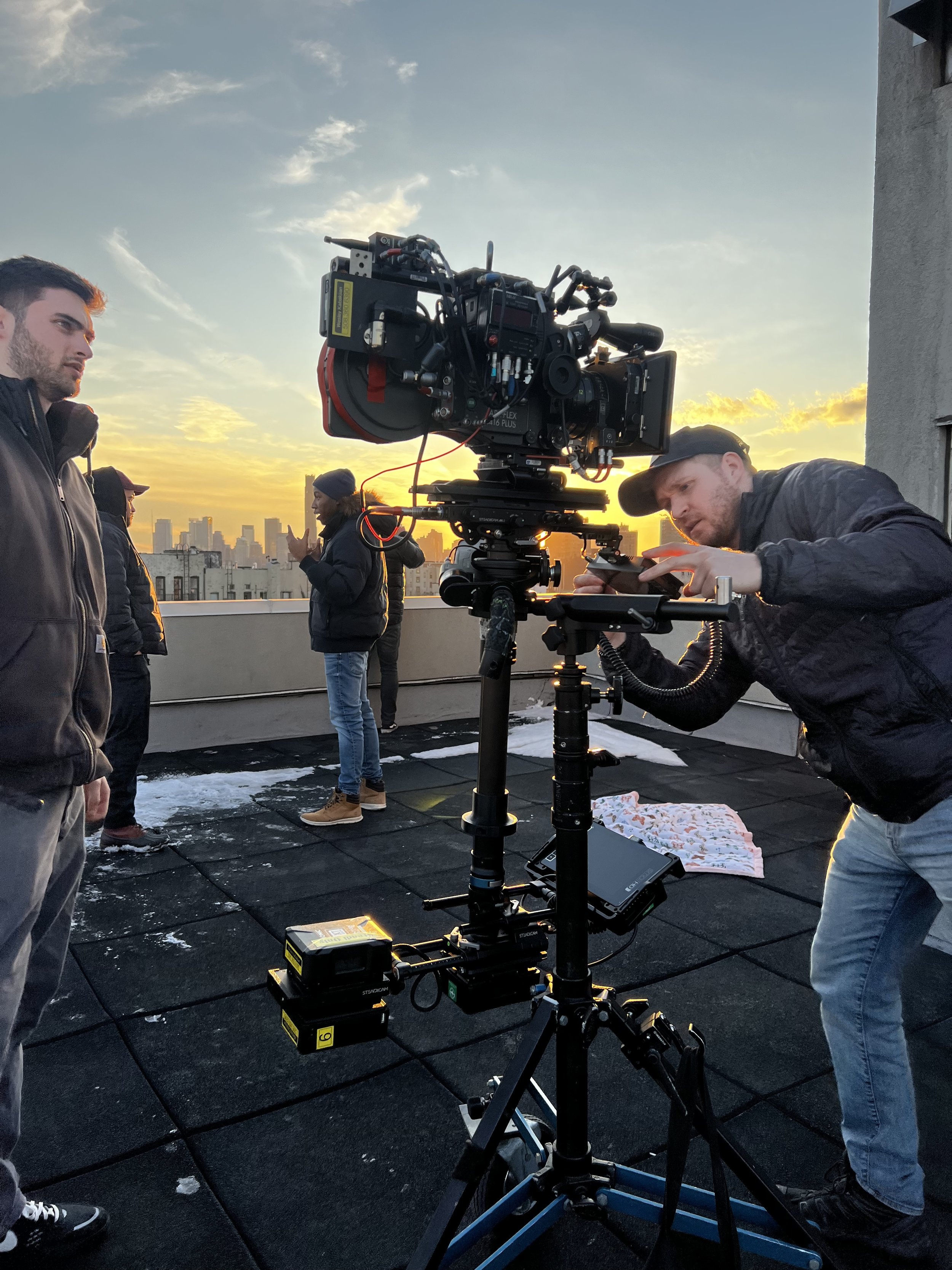 A film crew working on a rooftop during sunset, with a large camera mounted on a tripod and people in the background.
