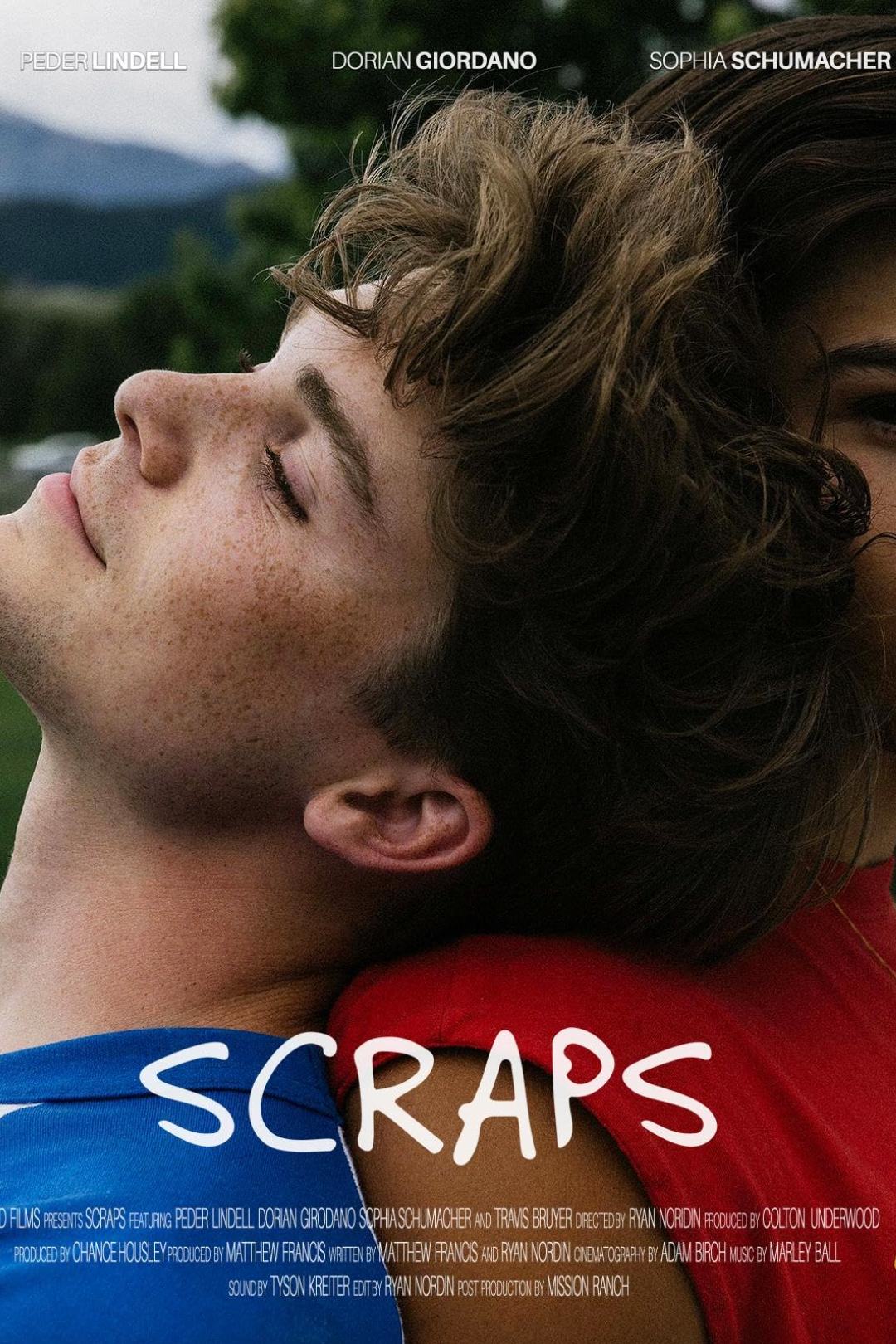 Close-up of two young people, one male with freckles and closed eyes with a content expression, and one female partially visible. They are resting with their heads touching outdoors, with a mountain and trees in the background. The text on the image 