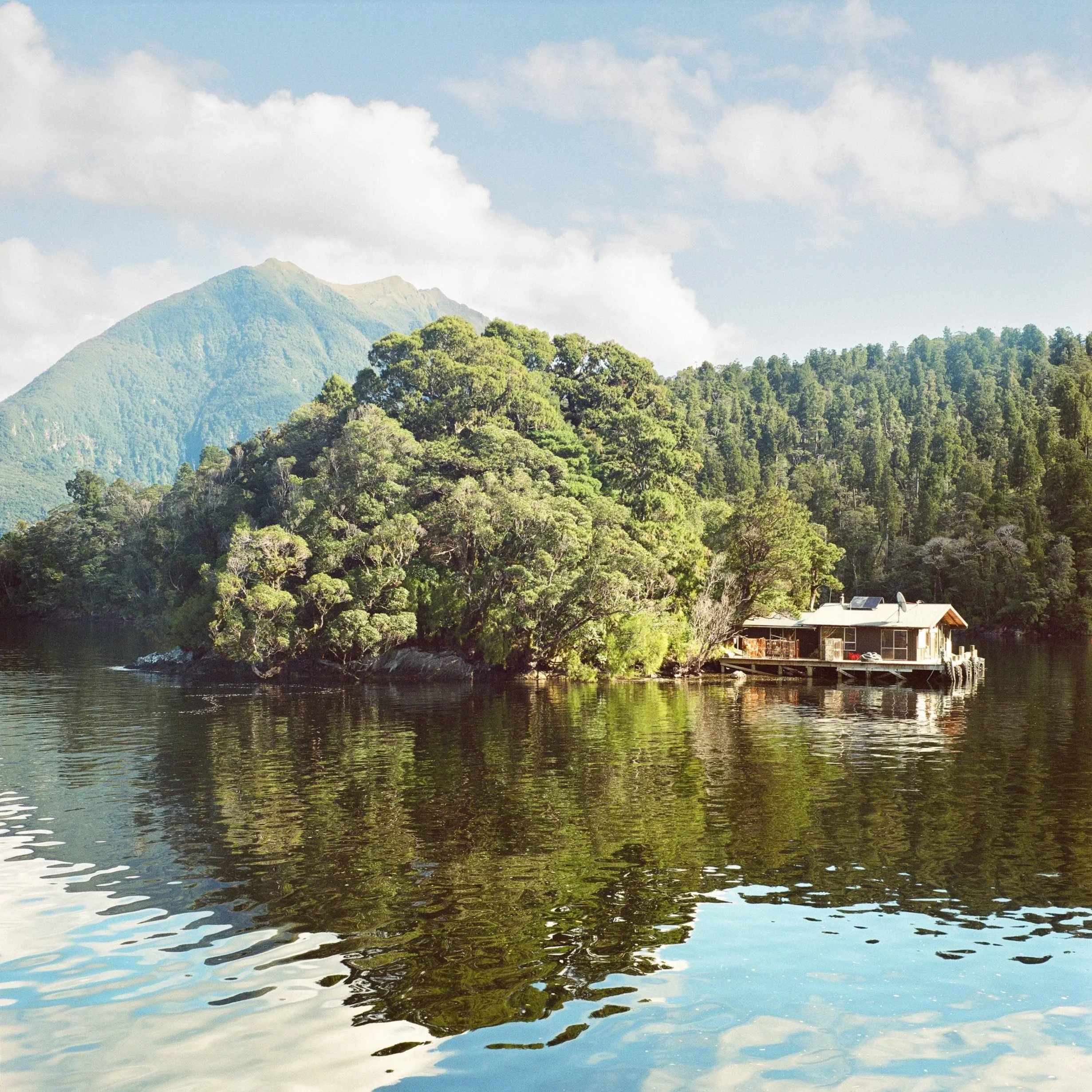 A house built on a boat anchored near a lush green island, surrounded by calm water with mountains in the background under a partly cloudy sky.
