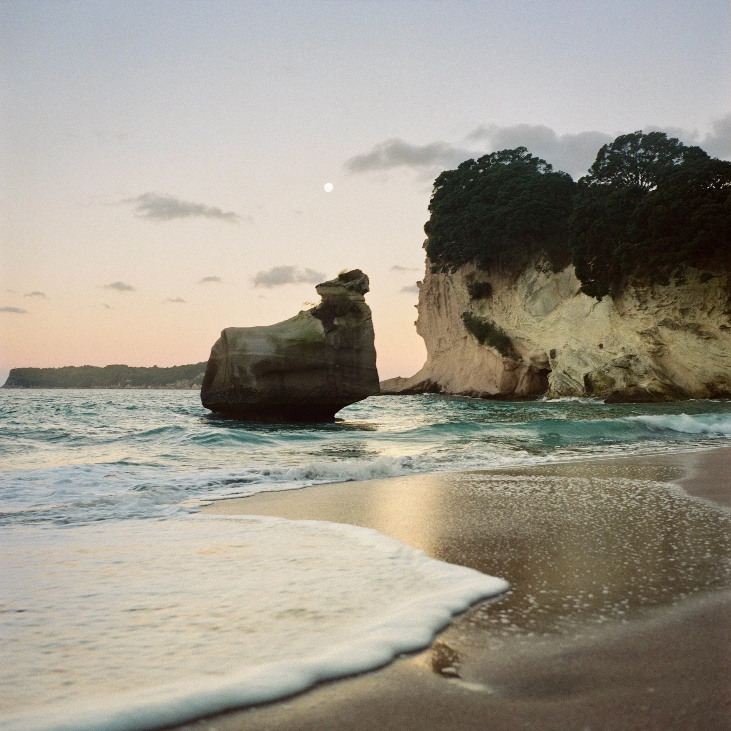 A coastal scene at sunset with a large rock formation in the ocean near a sandy beach, with cliffs and green trees in the background and the moon in the sky.