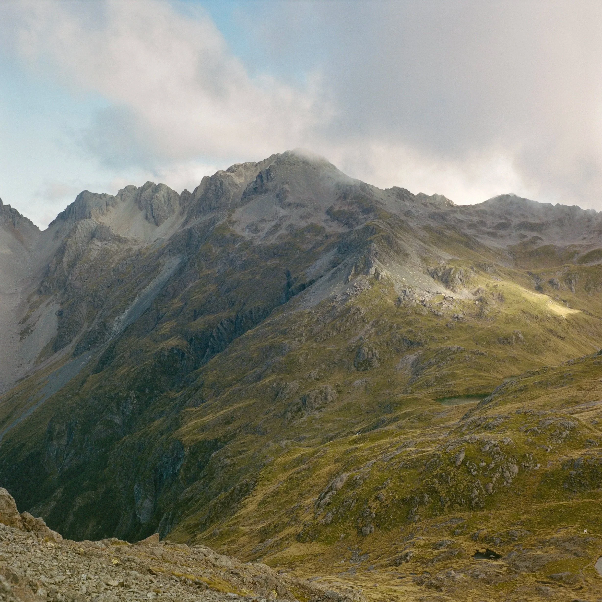 A mountain landscape with rugged peaks, partly covered in clouds, and green and rocky slopes.