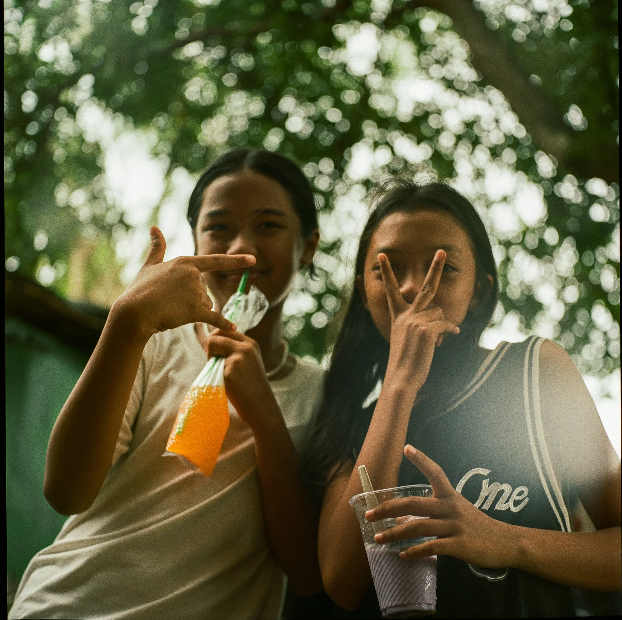 Two young women posing playfully outdoors, one holding an orange drink and the other holding a purple drink, under a leafy tree.